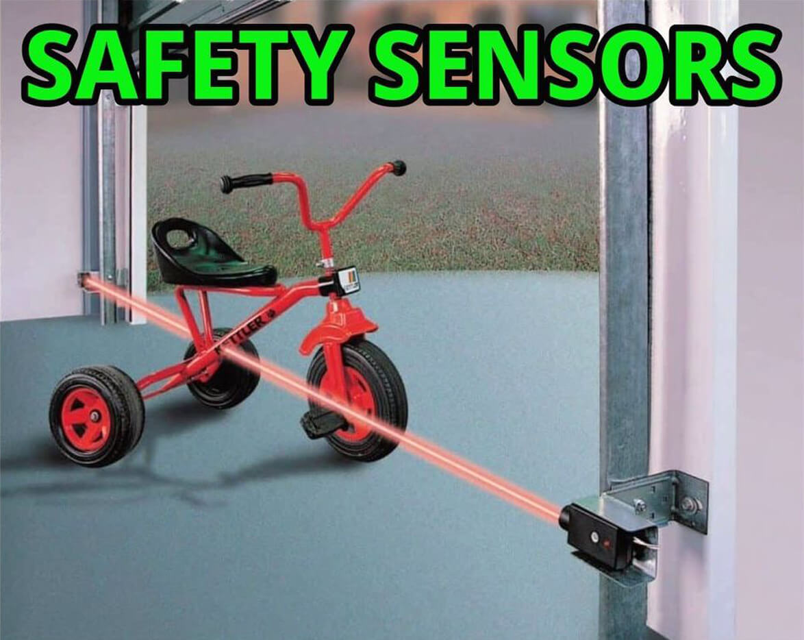 A red child's tricycle sits in the path of a garage door safety sensor's infrared beam, illustrating how the sensor works to prevent the door from closing on an obstruction.