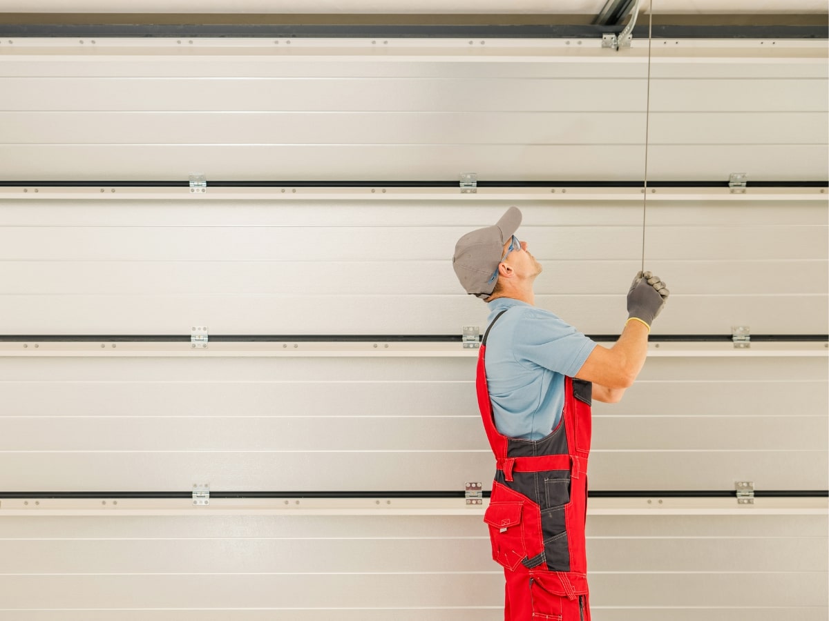 A technician in red overalls and a cap inspecting a garage door.